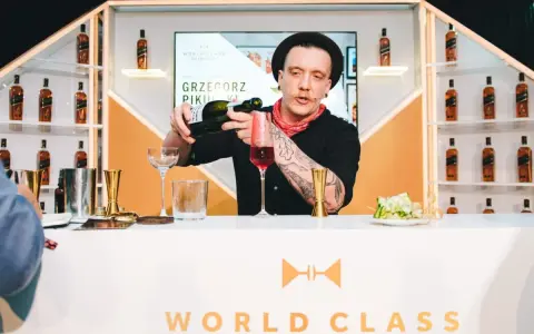 Barman Grzegorz Pikuła, w stylowym kapeluszu, nalewa drinka. W tle półki z butelkami whisky, idealne dla miłośników krwawych mary.
