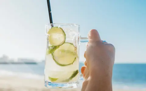 Orzeźwiająca Caipirinha czy Caipiroska z limonką i lodem, trzymana na tle plaży i błękitnego nieba. Idealny napój na wakacje.