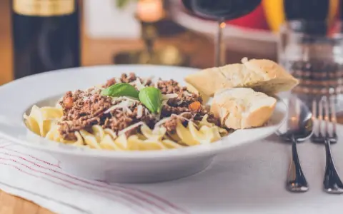 Pyszny makaron z sosem bolognese, posypany parmezanem i udekorowany bazylią. Czy to spaghetti, czy tagliatelle? Obok kromki chleba i kieliszek czerwonego wina.