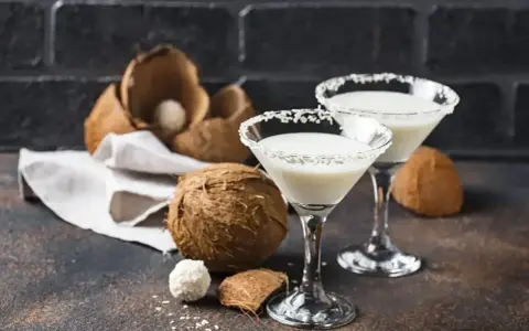Dwa kieliszki drinka z malibu z mlekiem, ozdobione wiórkami kokosowymi, obok świeżych kokosów.