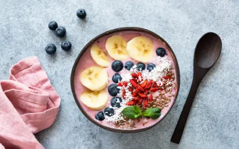 Kolorowy smoothie bowl z bananem, jagodami i kokosem, przypominający wyglądem tradycyjny kisiel. Obok leży drewniana łyżka i różowa serwetka.