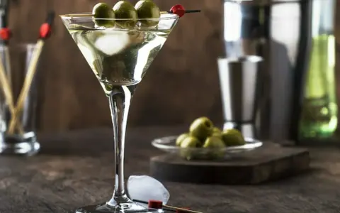 Elegancki drink z martini bianco, ozdobiony trzema oliwkami na patyczku. Obok kostki lodu i talerzyk z oliwkami.