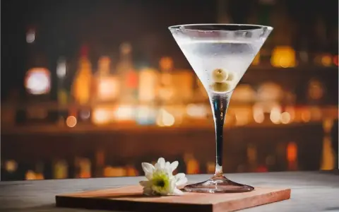 Eleganckie martini z colą, ozdobione oliwkami, na tle rozmytego baru. Obok kieliszka leży biały kwiatek.