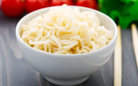 Biały makaron w misce, przypominający makaron konjac lub makaron z alg, z pomidorami i zielonymi warzywami w tle.