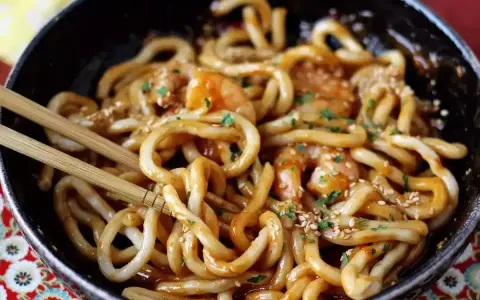 Miseczka z makaronem udon z krewetkami, posypana sezamem i pietruszką. Pałeczki zanurzone w daniu. Prosty przepis na to, jak gotować makaron udon.