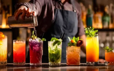 Barman przelewa drinka z shakera. Na blacie stoi rząd kolorowych koktajli, każdy ozdobiony owocami i miętą. Idealny przykład, jak zrobić dobry drink.
