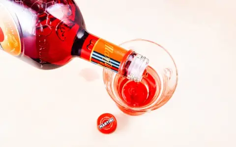 Czerwony Martini z sokiem pomarańczowym nalewany do kieliszka. Obok leży czerwona zakrętka z logo Martini.