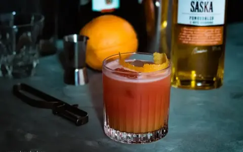 Orzeźwiający drink z likierem pomarańczowym, ozdobiony skórką cytrusową. Obok pomarańcza i butelka Saska.
