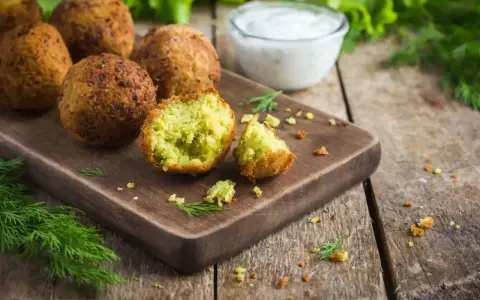 Chrupiące falafelki na desce, jeden przekrojony, ukazujący zielone wnętrze. Obok miseczka sosu i świeży koperek. Idealny falafel przepis.