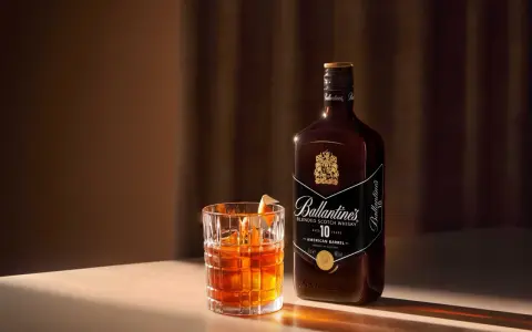 Whiskey z colą kontra Whiskey z ginger ale. Szklanka z bursztynowym trunkiem i butelka Ballantine's 10 Years Old.