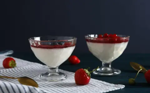 Dwie panna cotta: jedna na śmietance z borówkami, druga na jogurcie z malinami. Obok świeże truskawki i złote łyżeczki.