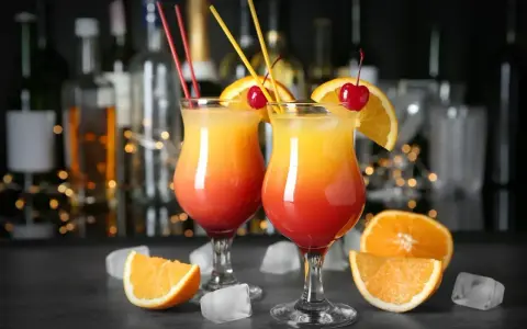 Dwa drinki Malibu z sokiem pomarańczowym, ozdobione pomarańczą i wiśnią, na tle baru.