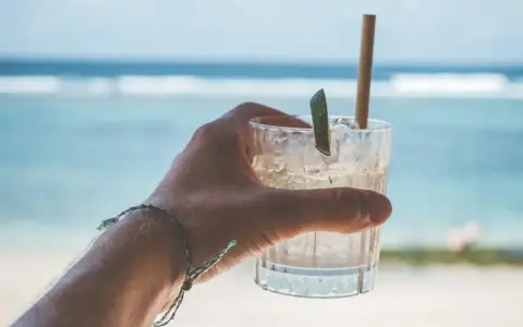 Orzeźwiający drink na plaży, z lodem i plasterkiem limonki, w dłoni z bransoletką. W tle błękitne morze i fale.