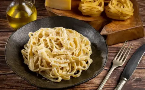 Pyszne Fettuccine Alfredo przepis na talerzu, obok surowy makaron, ser i oliwa. Idealne danie dla miłośników włoskiej kuchni.