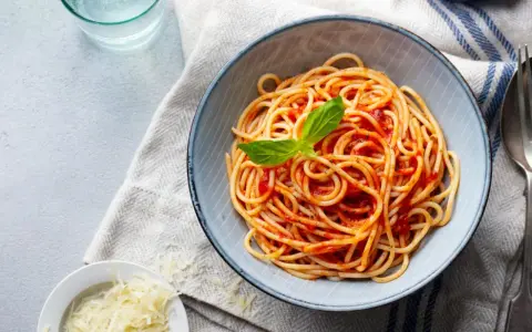 Miseczka spaghetti nr 5 z sosem pomidorowym i listkiem bazylii, obok spaghettini, starty parmezan i szklanka wody.