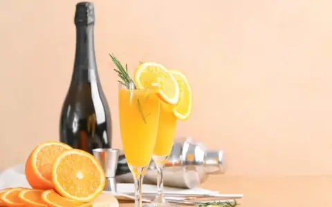 Dwa kieliszki z pomarańczowym napojem, ozdobione plasterkami pomarańczy i rozmarynem. Obok butelka szampana, shaker i świeże pomarańcze. Mimosa czy Bellini?