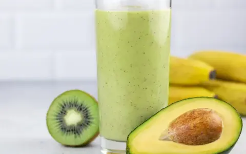 Zielony, kremowy koktajl z awokado, kiwi i banana. Obok szklanki połówka awokado i przekrojone kiwi. W tle pęk bananów.