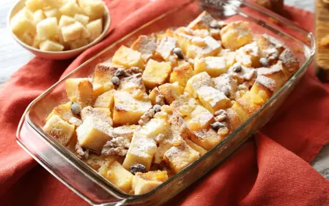 Pudding chlebowy w naczyniu, posypany cukrem pudrem i rodzynkami, obok miseczka z pokrojonym chlebem.