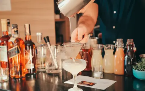 Barman przelewa płyn z shakera przez sitko do kieliszka, tworząc koktajl. W tle butelki alkoholi i kolorowe syropy.