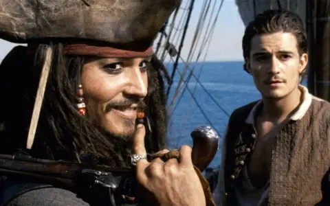 Jack Sparrow, z fajką i karabinem, uśmiecha się do Willa Turnera. Pewnie myśli o kolejnej butelce rumu.