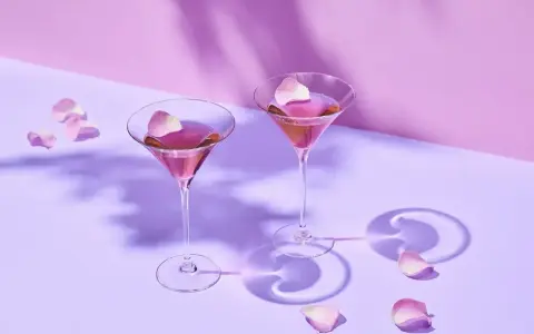 Elegancki Martini przepis: dwa kieliszki z różowym drinkiem, ozdobione płatkami róż, na fioletowym tle.
