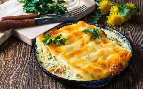 Pyszne conchiglioni zapieczone z serem i ziołami, podane w czarnej patelni. Obok deska z nożem i widelcem, oraz pęczek natki pietruszki.
