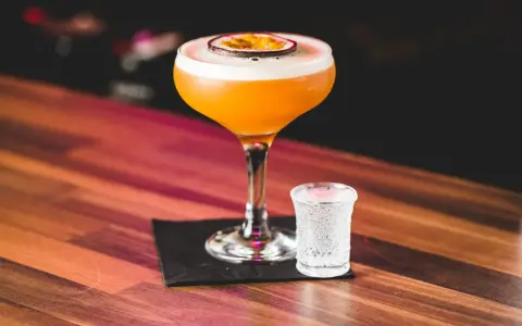 Orzeźwiający drink z marakują, ozdobiony plasterkiem owocu, stoi obok kieliszka z wodą gazowaną na drewnianym blacie.