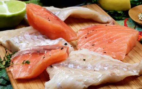Stek z łososia kontra polędwica z dorsza. Surowe kawałki ryb, przyprawione pieprzem, leżą obok limonki, cytryny i ziół.