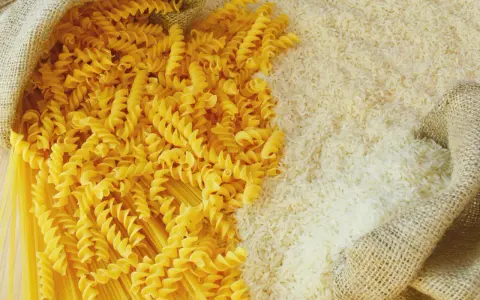 Ryż vs makaron: obok siebie leżą dwa rodzaje produktów. Z worka wysypano biały ryż, a obok niego spiralne fusilli i długie spaghetti.