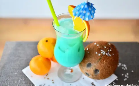 Orzeźwiający drink z sokiem ananasowym, ozdobiony plasterkiem pomarańczy i parasolką. Obok leżą pomarańcze i kokos.
