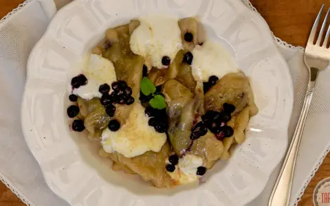Knedle ze śliwkami czy pierogi z jagodami? Na talerzu pyszne pierogi z jagodami, polane śmietaną i posypane świeżymi owocami.