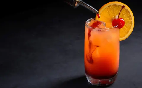 Kolorowy drink z syropem grenadina, ozdobiony plasterkiem pomarańczy i wisienką.