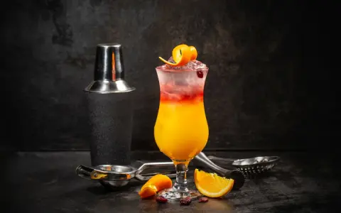 Kolorowy drink z lodem, pomarańczą i żurawiną, obok shakera. Wybór między Cosmopolitan a Sex on the Beach.