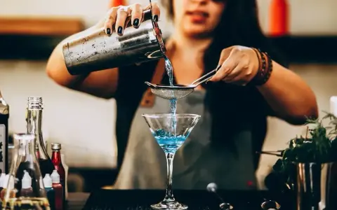 Barman przygotowuje drinka, przelewając niebieski płyn przez sitko do kieliszka. W tle widać butelki z różnymi nazwami drinków alkoholowych.