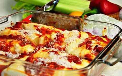 Pyszne, zapiekane cannelloni w sosie pomidorowym, posypane parmezanem. Obok świeże warzywa, idealne do dania.
