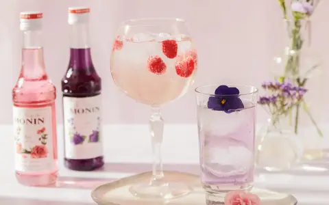 Dobre drinki przepisy: różowy i fioletowy syrop Monin, drink z malinami w kielichu, drink z fiołkiem w szklance, kwiaty w wazonach.
