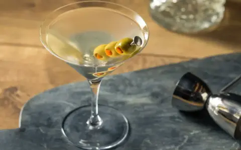 Elegancki martini z wódką, ozdobiony trzema oliwkami na wykałaczce, gotowy do degustacji. Obok metalowy jigger.