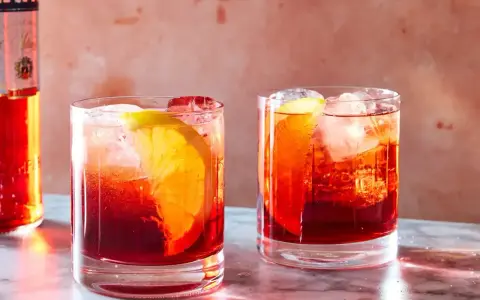 Dwa drinki Negroni z pomarańczą i lodem, obok butelki Campari. Idealny drink negroni przepis na lato.
