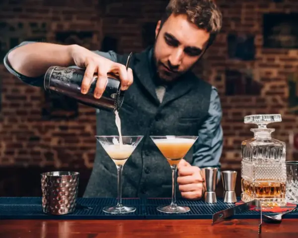 Barman przelewa drinka do kieliszka, pokazując, jak być dobrym barmanem. Na blacie stoją też inne drinki i akcesoria.