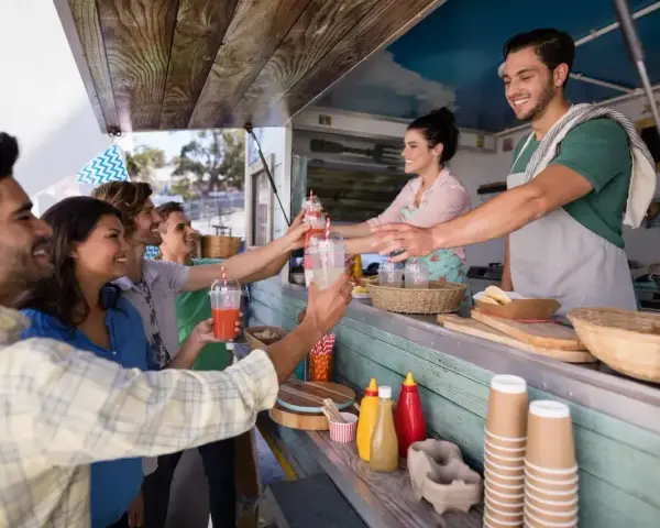 Uśmiechnięci klienci odbierają napoje od barmana w food trucku. Ciekawe, ile zarabia barman w takim miejscu?