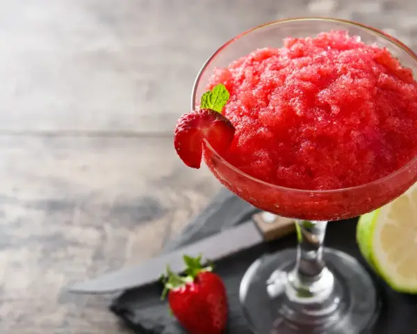 Orzeźwiający strawberry daiquiri, udekorowany plasterkiem truskawki i listkiem mięty, obok połówki limonki i świeżej truskawki.