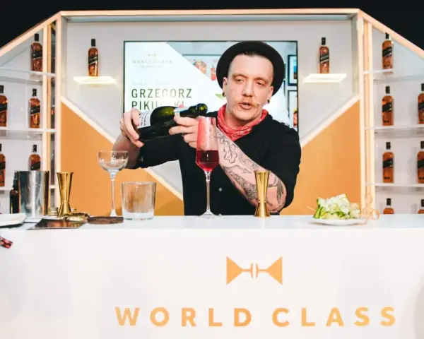 Barman Grzegorz Pikuła, w stylowym kapeluszu, nalewa drinka. W tle półki z butelkami whisky, idealne dla miłośników krwawych mary.