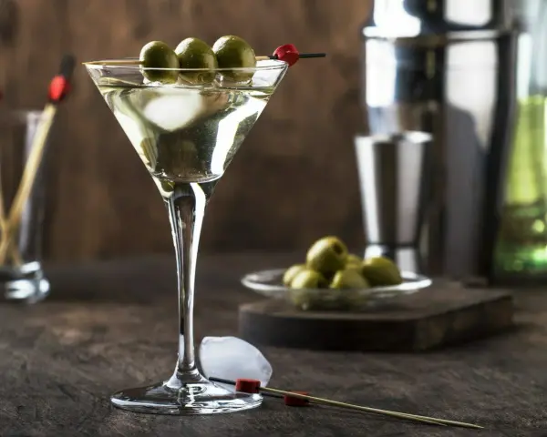 Elegancki drink z martini bianco, ozdobiony trzema oliwkami na patyczku. Obok kostki lodu i talerzyk z oliwkami.