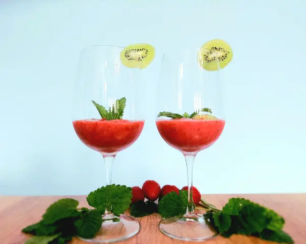 Dwa kieliszki orzeźwiającego drinka truskawkowego z rumem, ozdobione plasterkami kiwi i miętą. Na drewnianym blacie leżą świeże truskawki i listki mięty.
