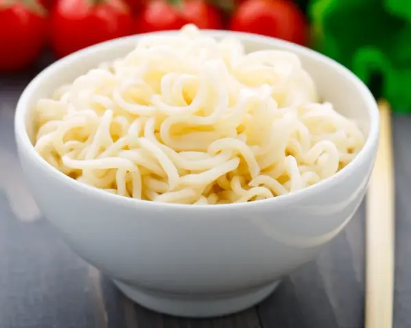 Biały makaron w misce, przypominający makaron konjac lub makaron z alg, z pomidorami i zielonymi warzywami w tle.