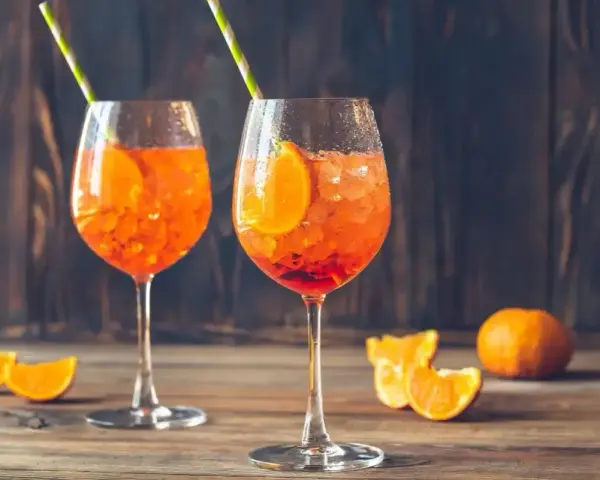 Dwa orzeźwiające drinki z aperolem, ozdobione plastrami pomarańczy i słomkami, na drewnianym blacie.