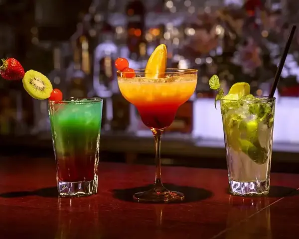Trzy kolorowe drinki na barze. Jeden z nich to drink z pomarańczą, ozdobiony owocami.