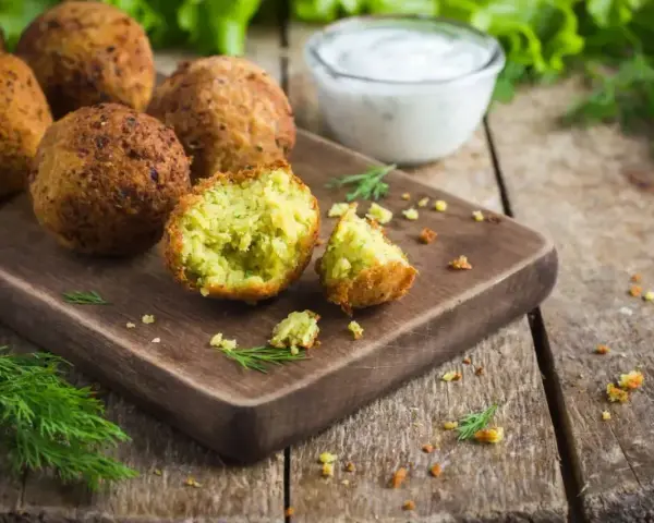 Chrupiące falafelki na desce, jeden przekrojony, ukazujący zielone wnętrze. Obok miseczka sosu i świeży koperek. Idealny falafel przepis.