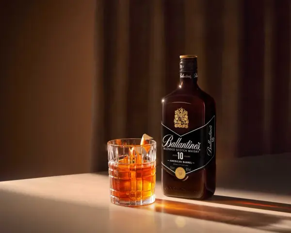 Whiskey z colą kontra Whiskey z ginger ale. Szklanka z bursztynowym trunkiem i butelka Ballantine's 10 Years Old.