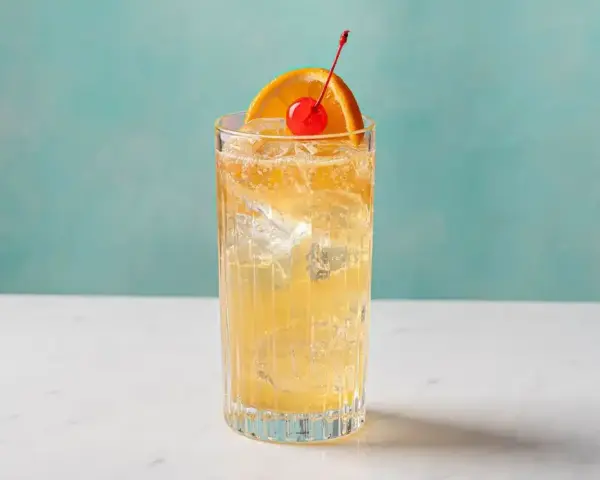 Orzeźwiający John Collins drink z lodem, plasterkiem pomarańczy i wisienką na tle turkusowej ściany.